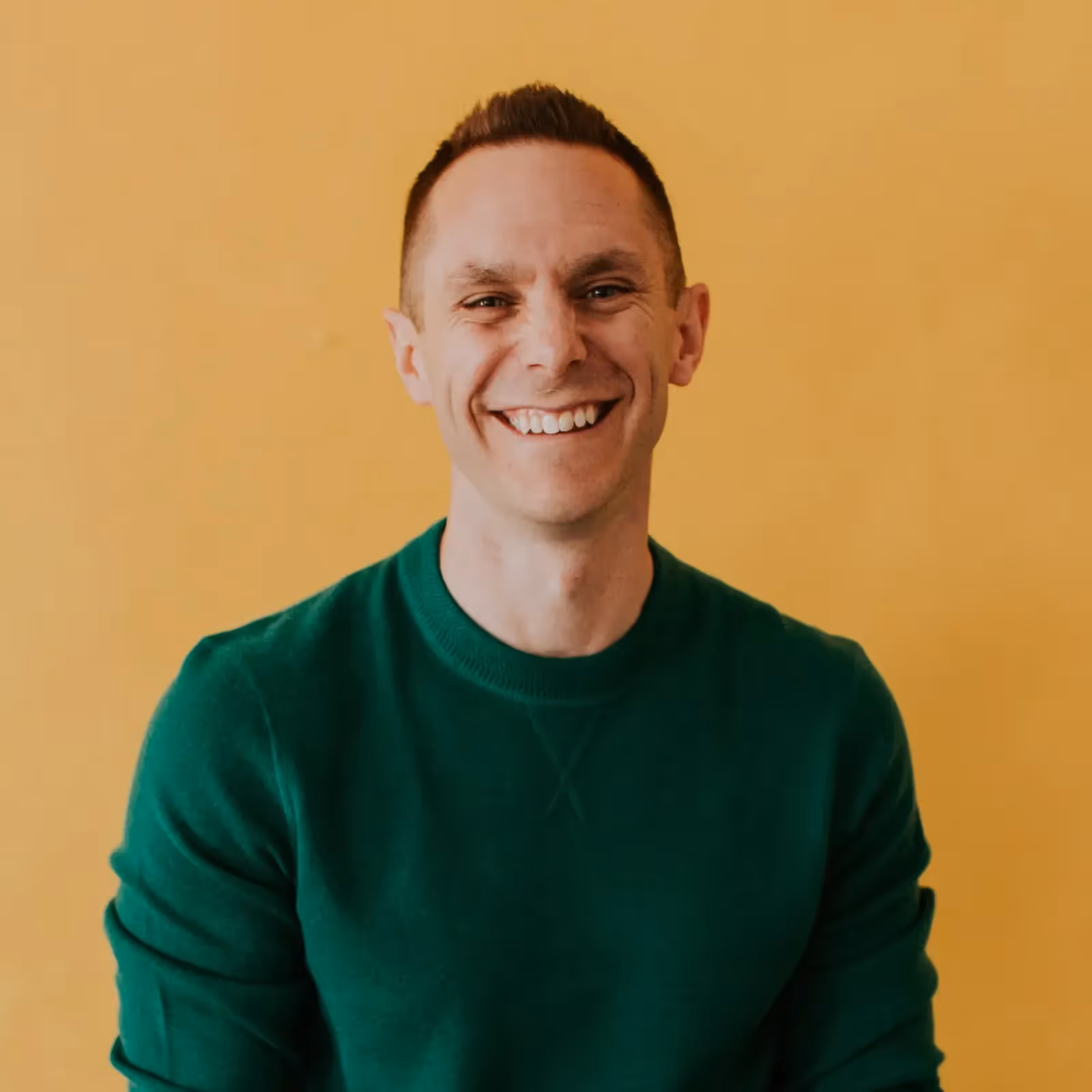 Smiling man in a green sweater against a yellow background.