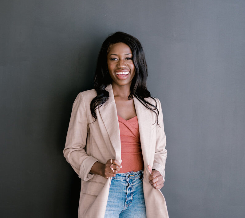 Portrait of a smiling woman in a beige blazer and jeans against a dark background.