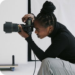 A woman with braided hair styled in a bun is crouched while looking through the viewfinder of a professional camera, preparing to take a photo.