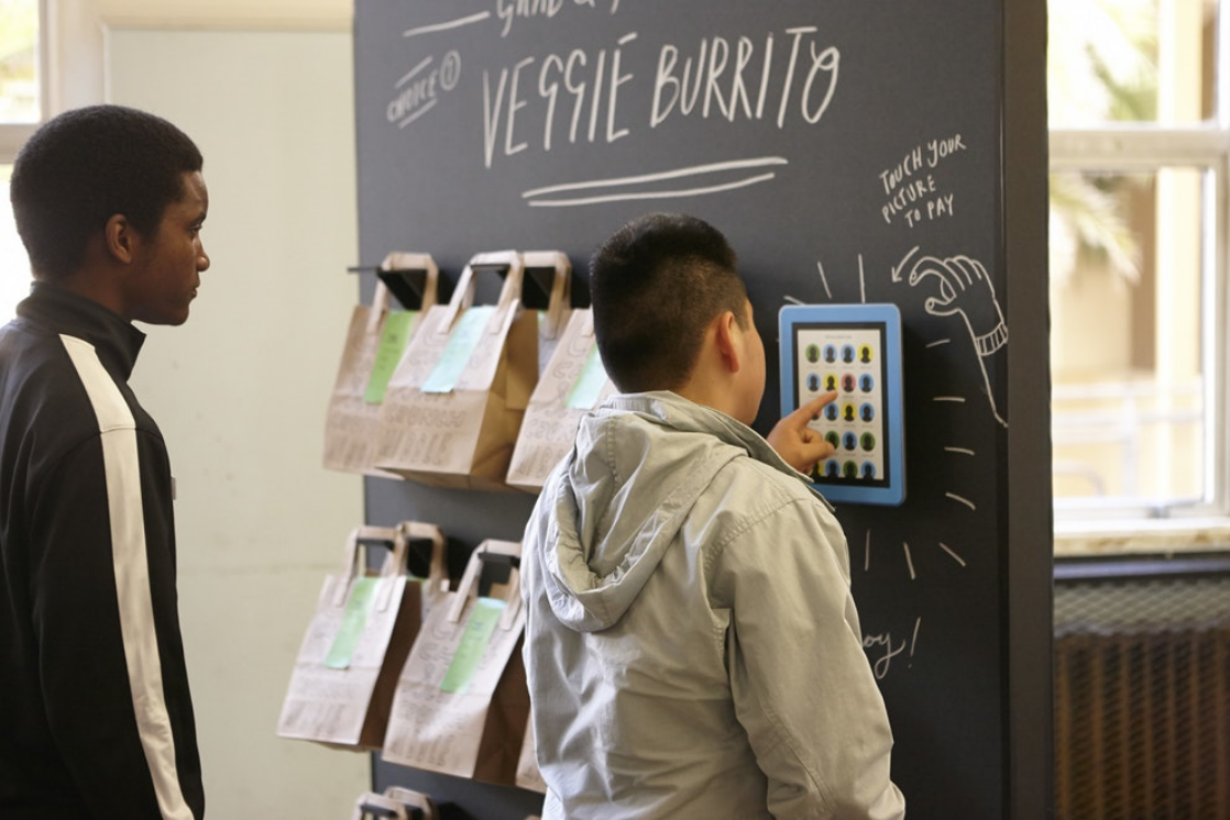 Students using a tablet kiosk to pay for a veggie burrito next to hanging paper lunch bags labeled with student names