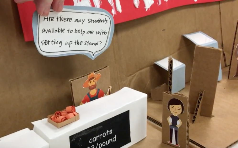 Low-fidelity paper prototype scene with a speech bubble asking if students are available to help set up the market stand