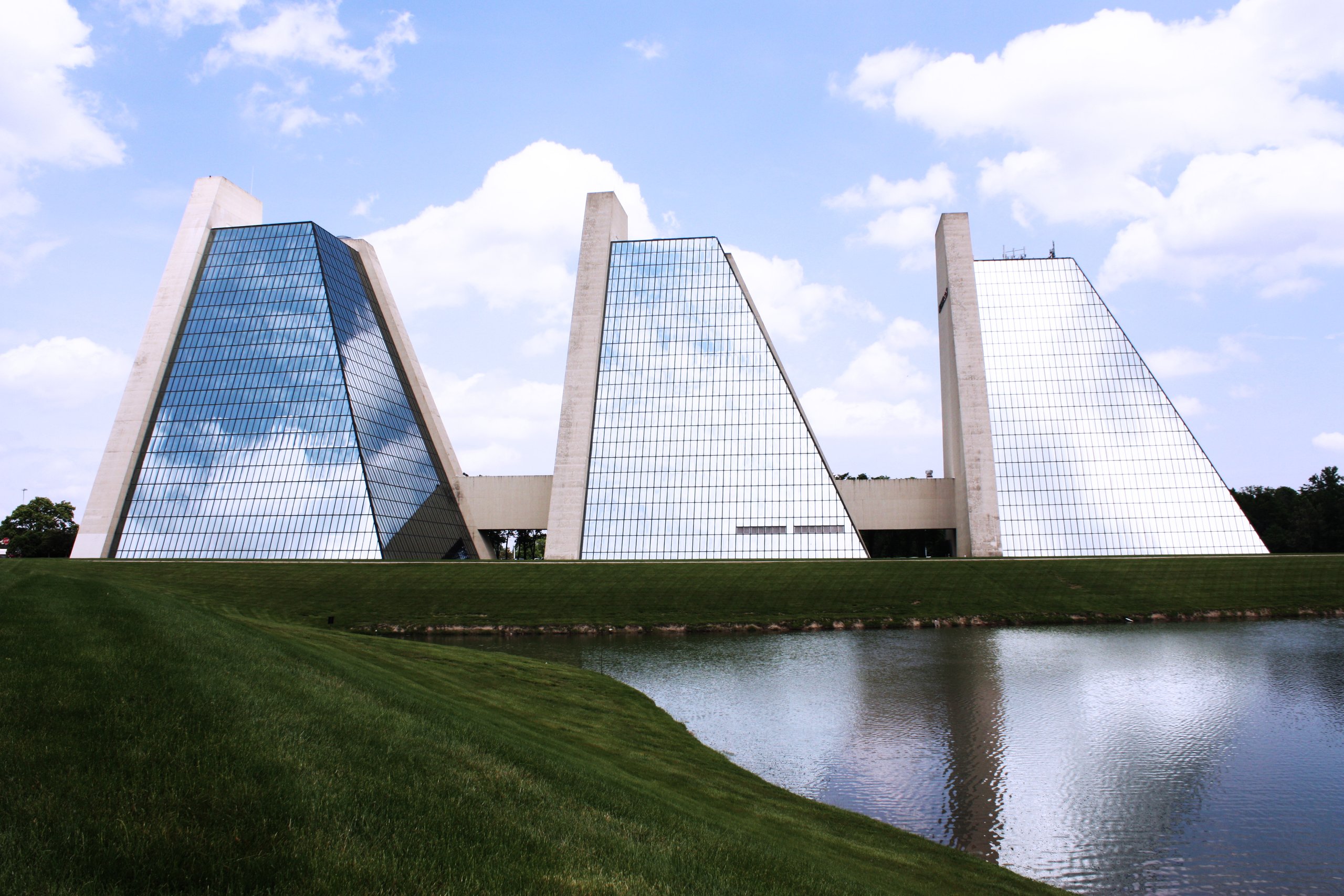 Three glass pyramid office buildings with tall concrete towers beside a pond and grassy bank under a partly cloudy sky