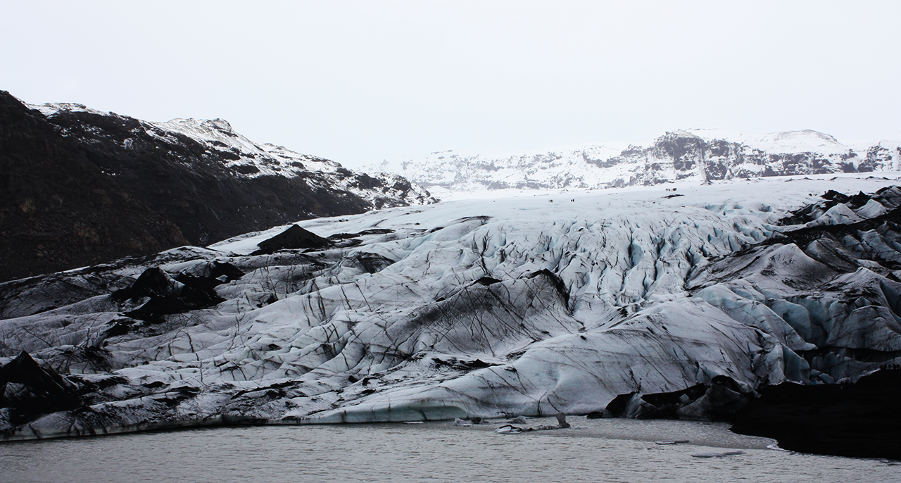 Glacier with ash-streaked ice spilling between dark mountains, with a gray lagoon in the foreground