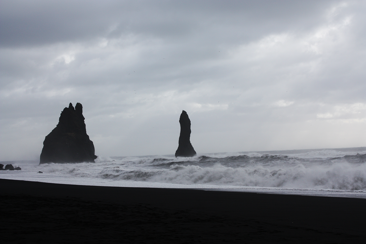 Waves breaking on a black sand beach in front of two tall rock sea stacks under a stormy sky