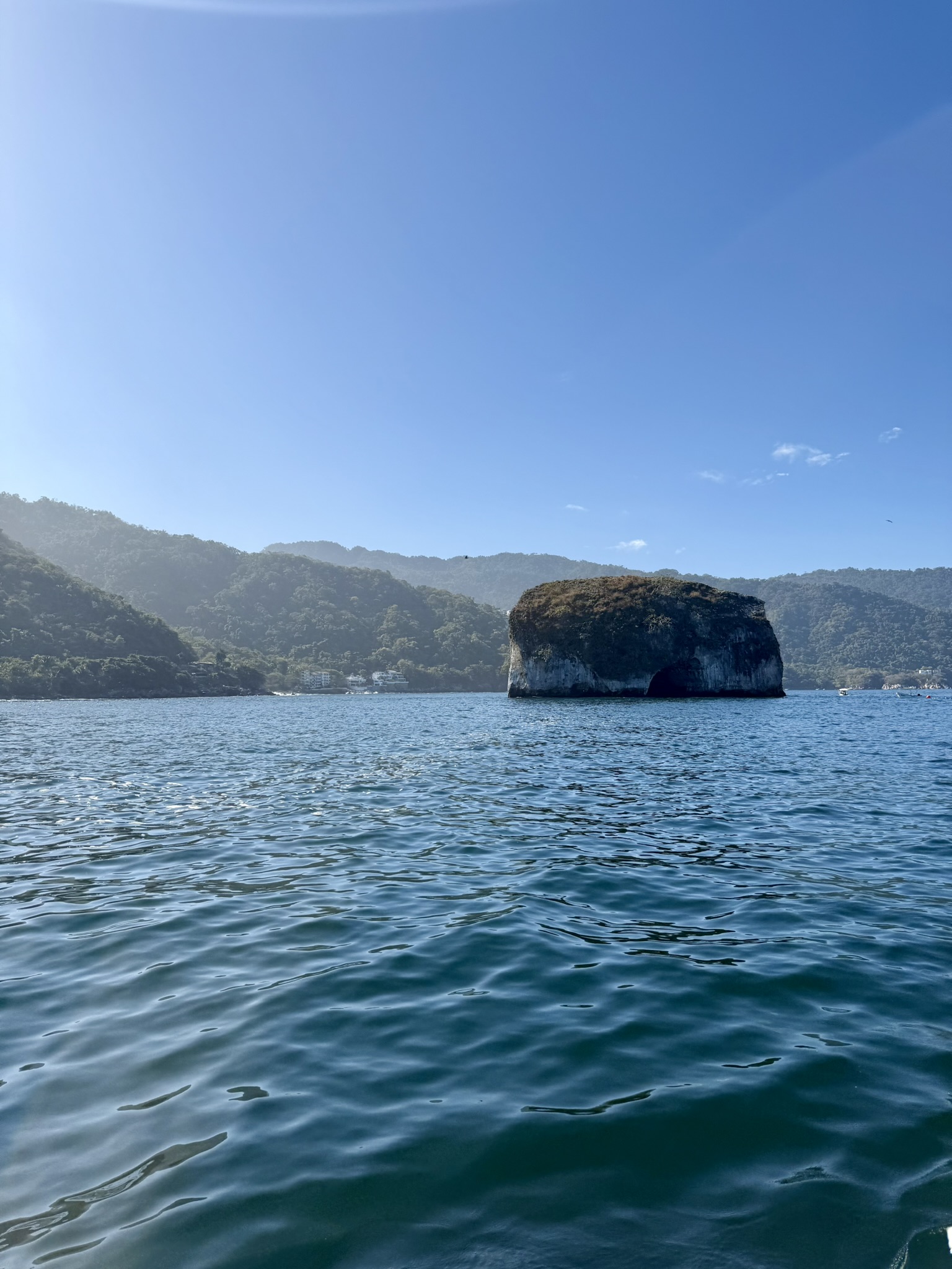 Rocky island rising from calm blue water in a bay surrounded by green hills