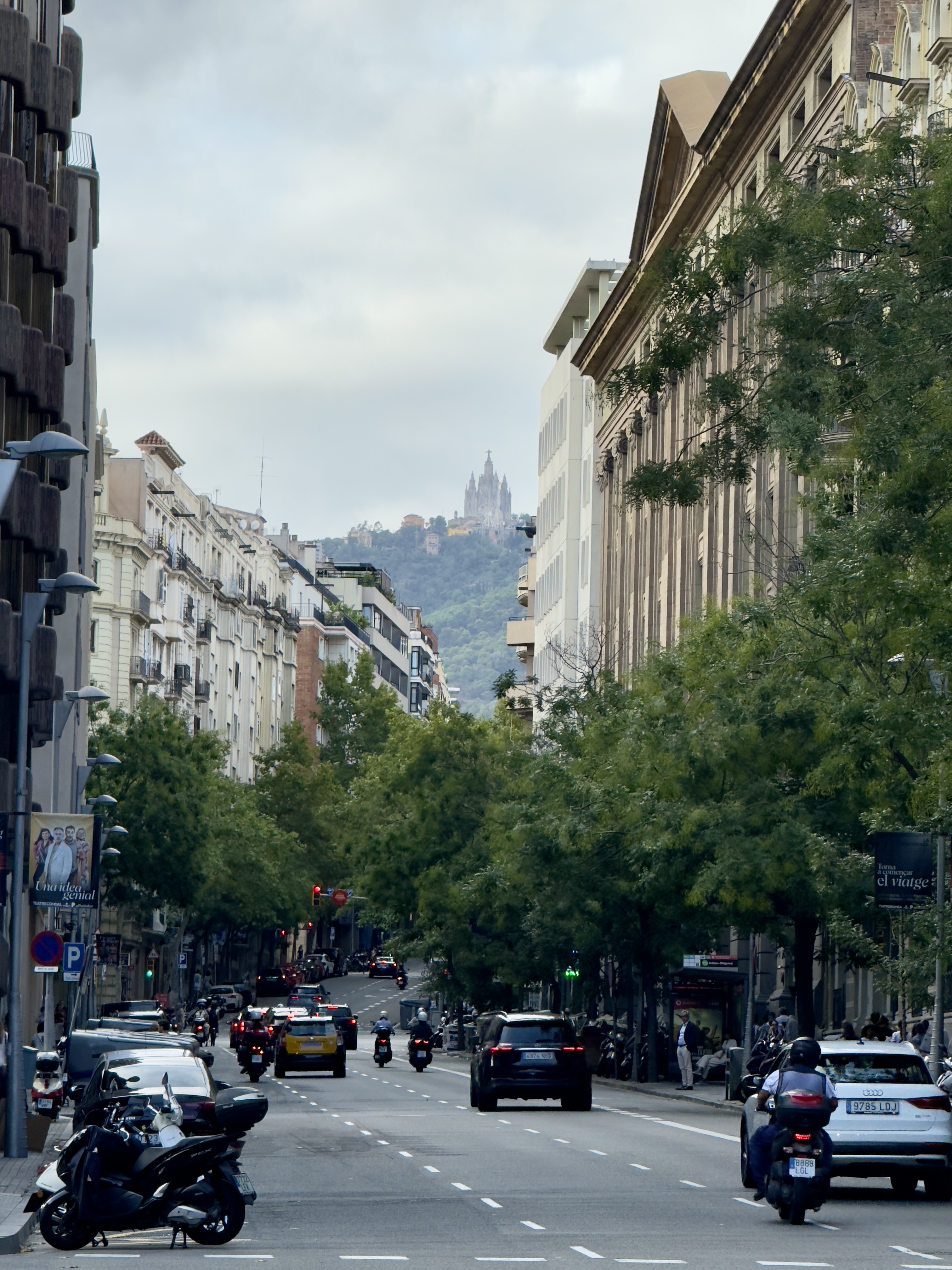 Tree-lined city avenue with cars and scooters, leading toward a church on a distant hilltop
