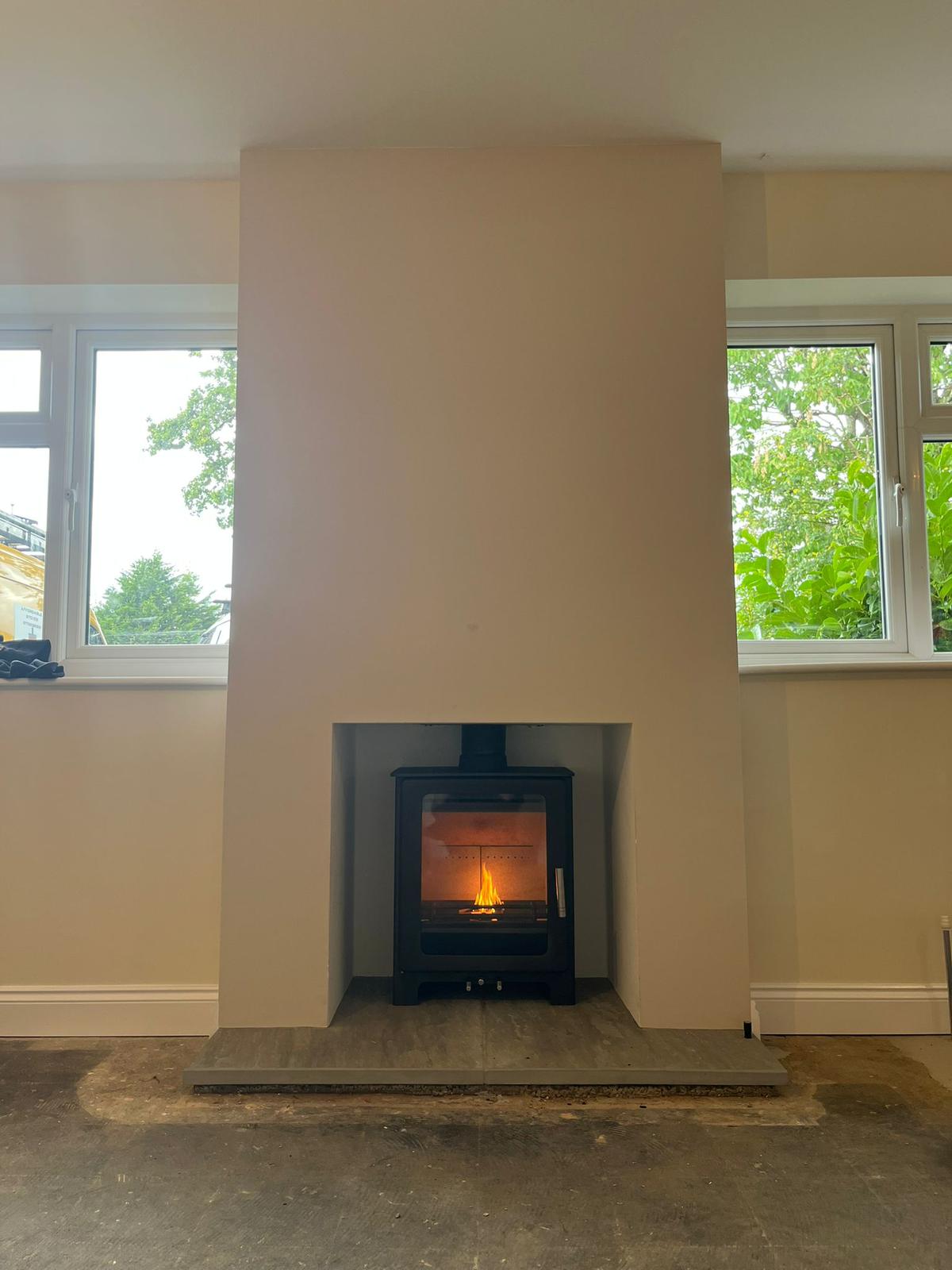 Modern black wood-burning stove with visible flame inside a beige fireplace recess, flanked by two windows showing green trees outside.