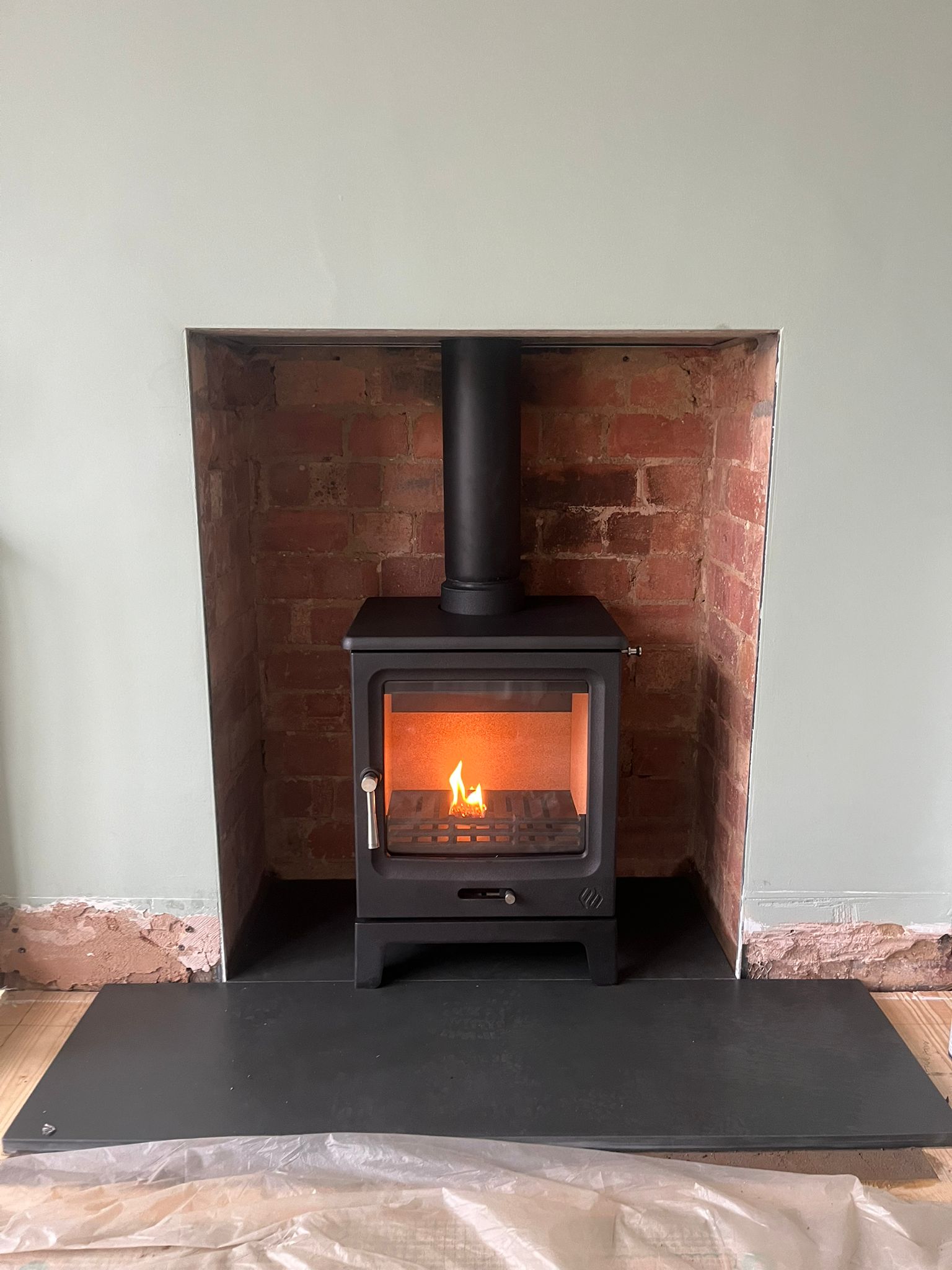 Modern black wood-burning stove lit with a small fire inside a red brick fireplace recess.