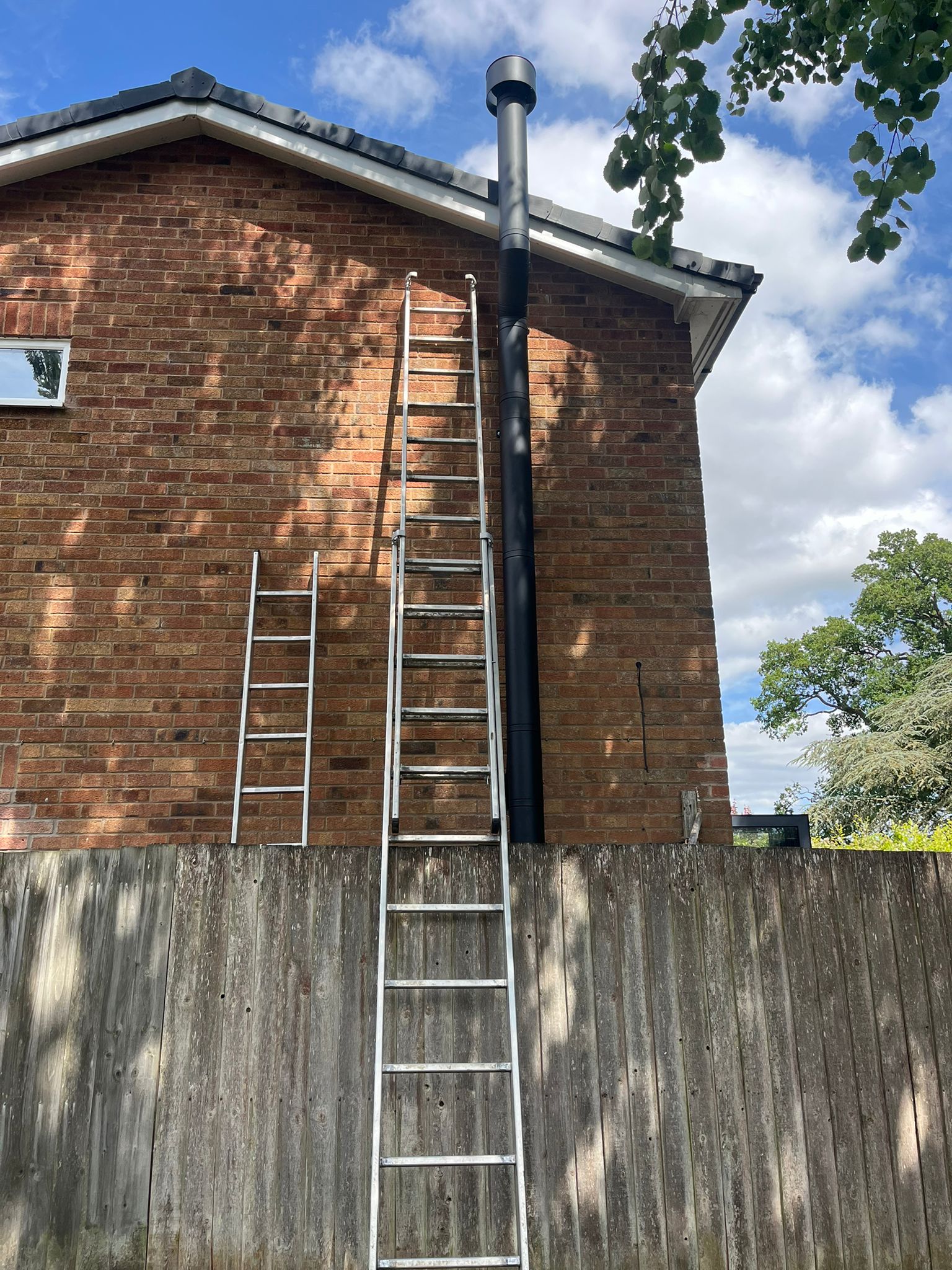 Two metal ladders leaning against a brick house with a tall black chimney pipe, wooden fence in the foreground, and trees with blue sky in the background.