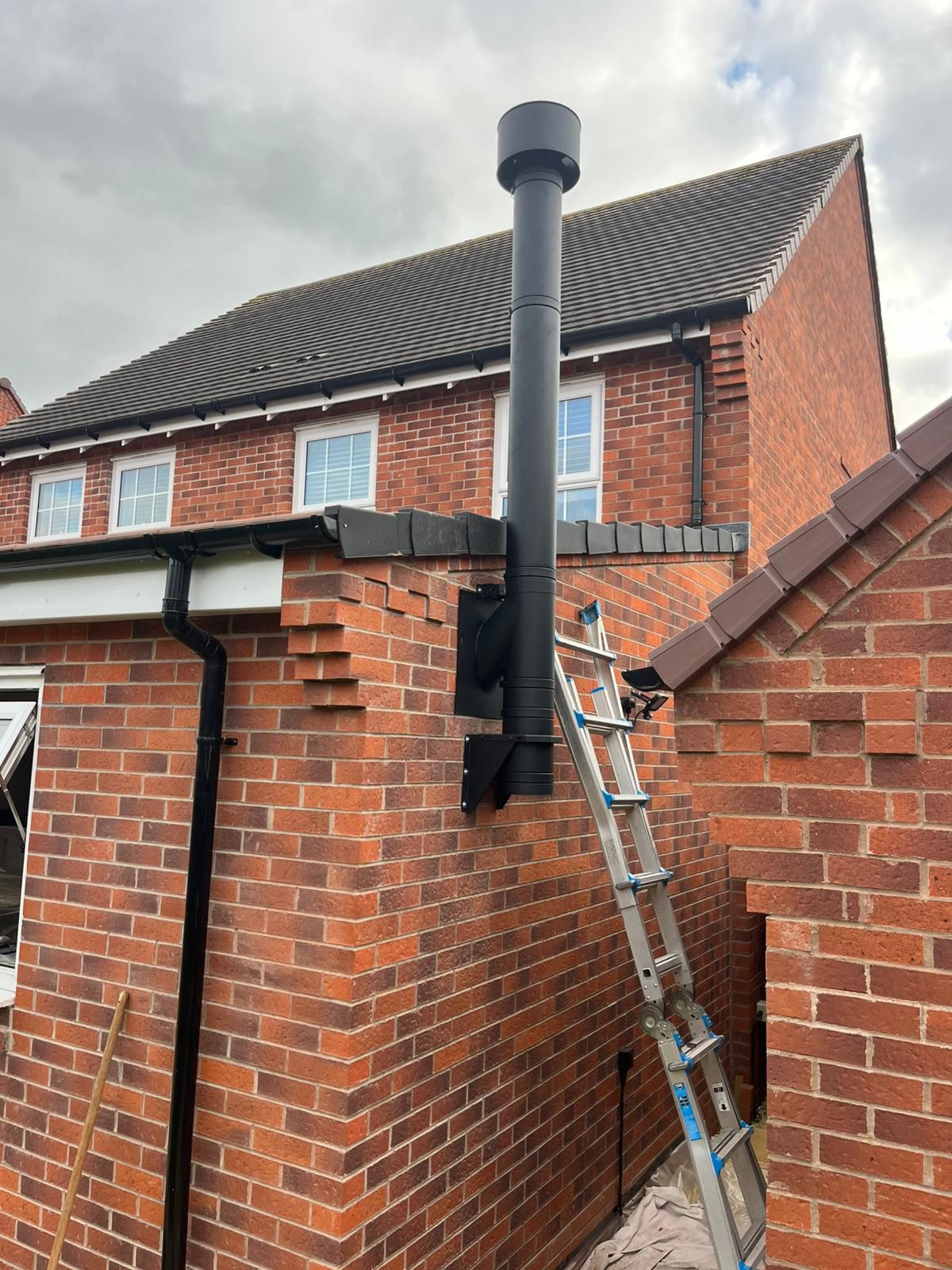Black metal chimney pipe installed on a red brick house exterior next to a metal ladder.