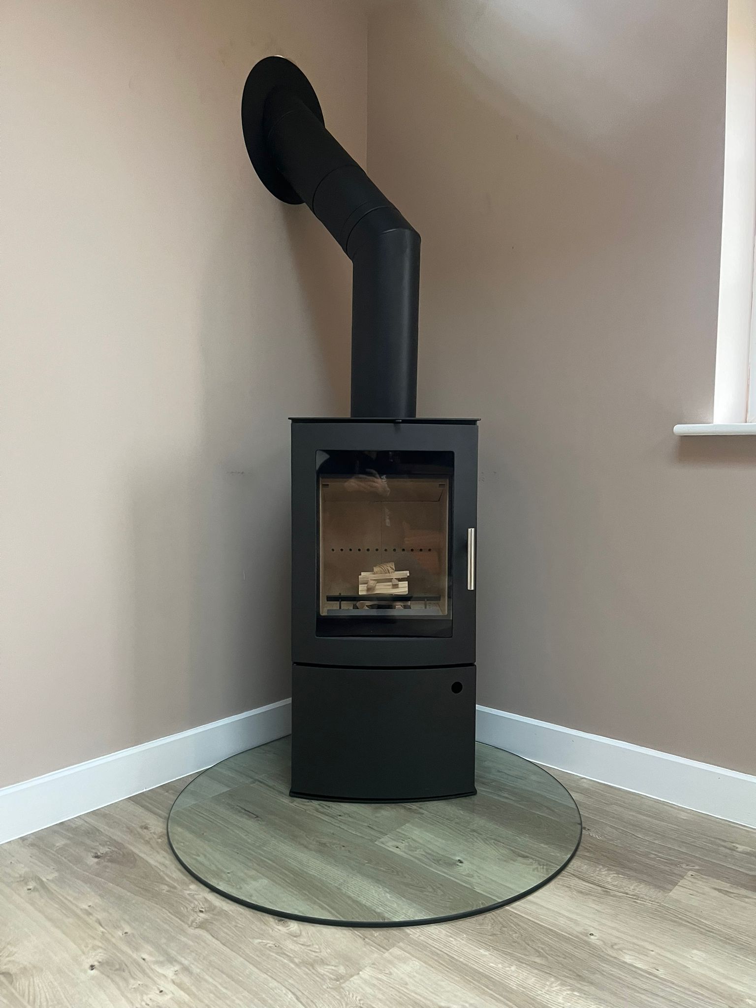Modern black wood-burning stove with glass door and chimney pipe installed in a beige corner on a circular glass hearth.