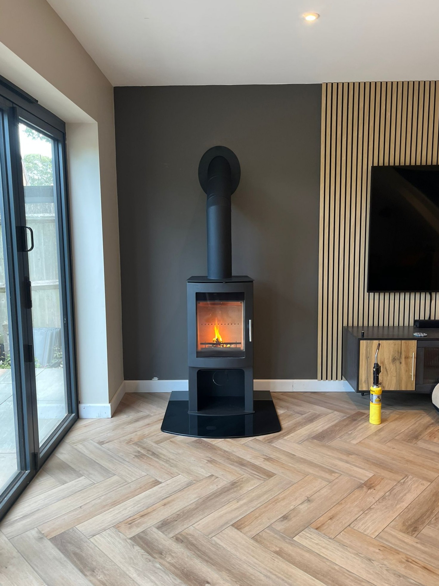 Modern wood-burning stove with fire inside against dark grey wall, next to wood paneled wall and TV, on herringbone wooden floor.