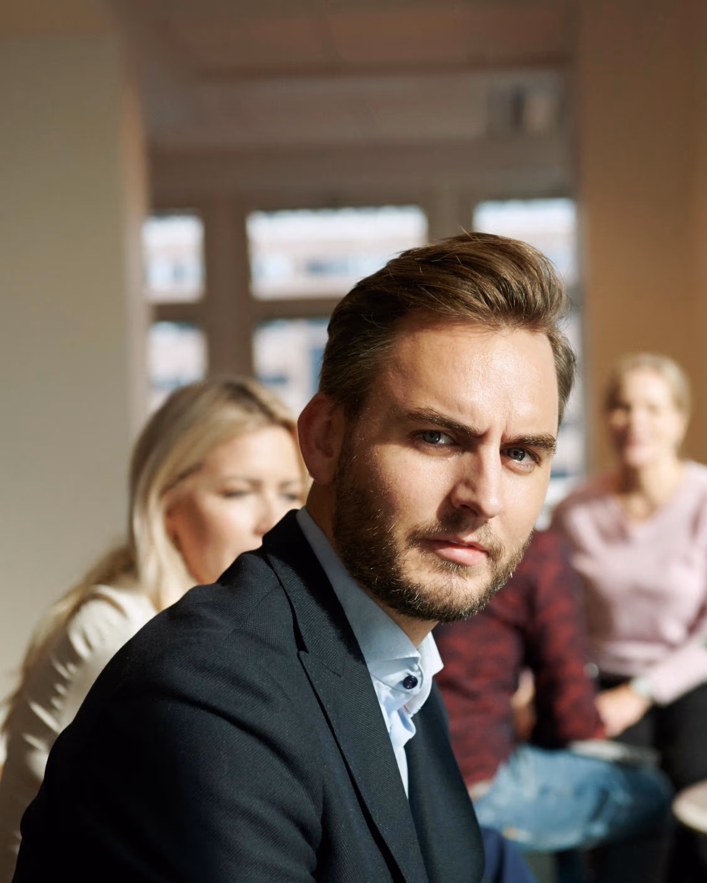 Nærbilde av en mann i dress som ser alvorlig mot kamera, med tre uskarpe personer bak.