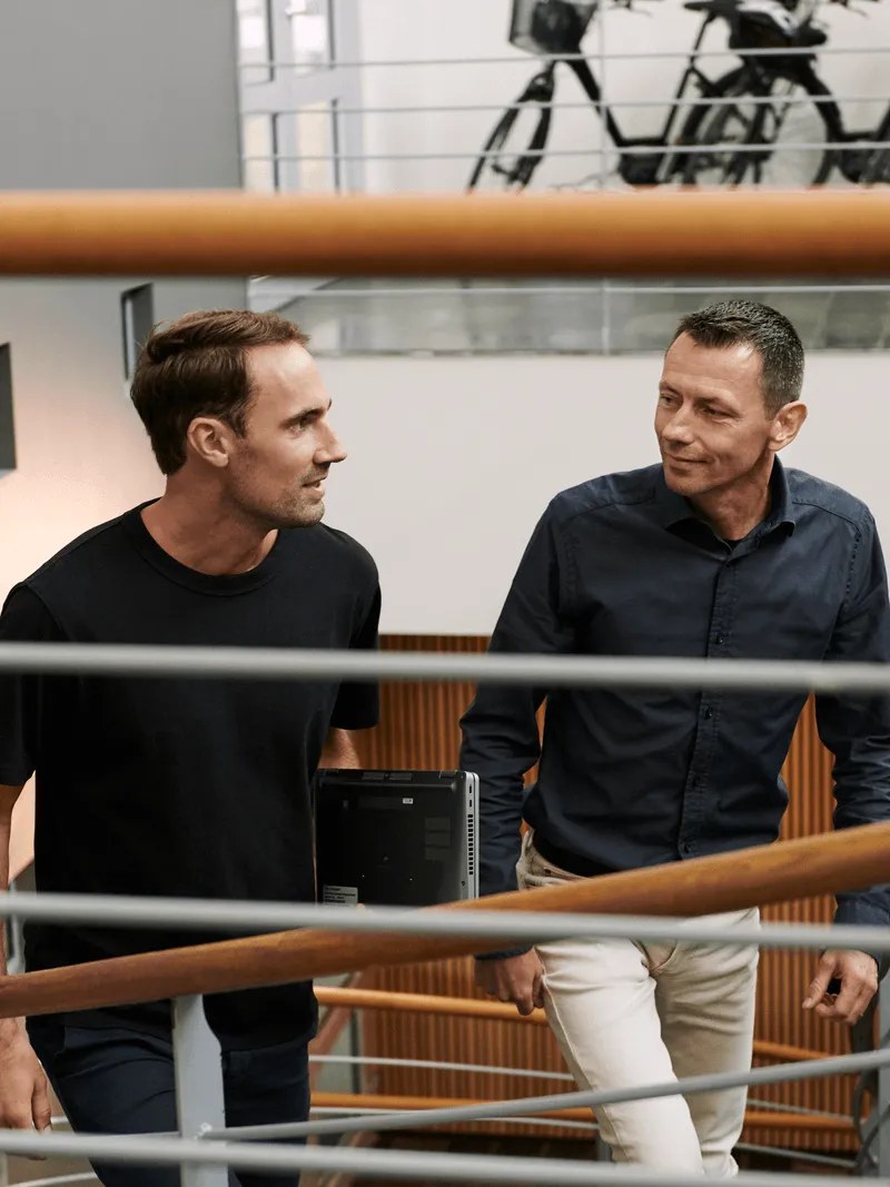 Bildet viser to menn som går opp en trapp i et moderne bygg; den ene mannen bærer en bærbar datamaskin og er kledd i en svart t-skjorte, mens den andre har på seg en mørk skjorte og lyse bukser. Bak dem kan man se sykler parkert på en mesanin.