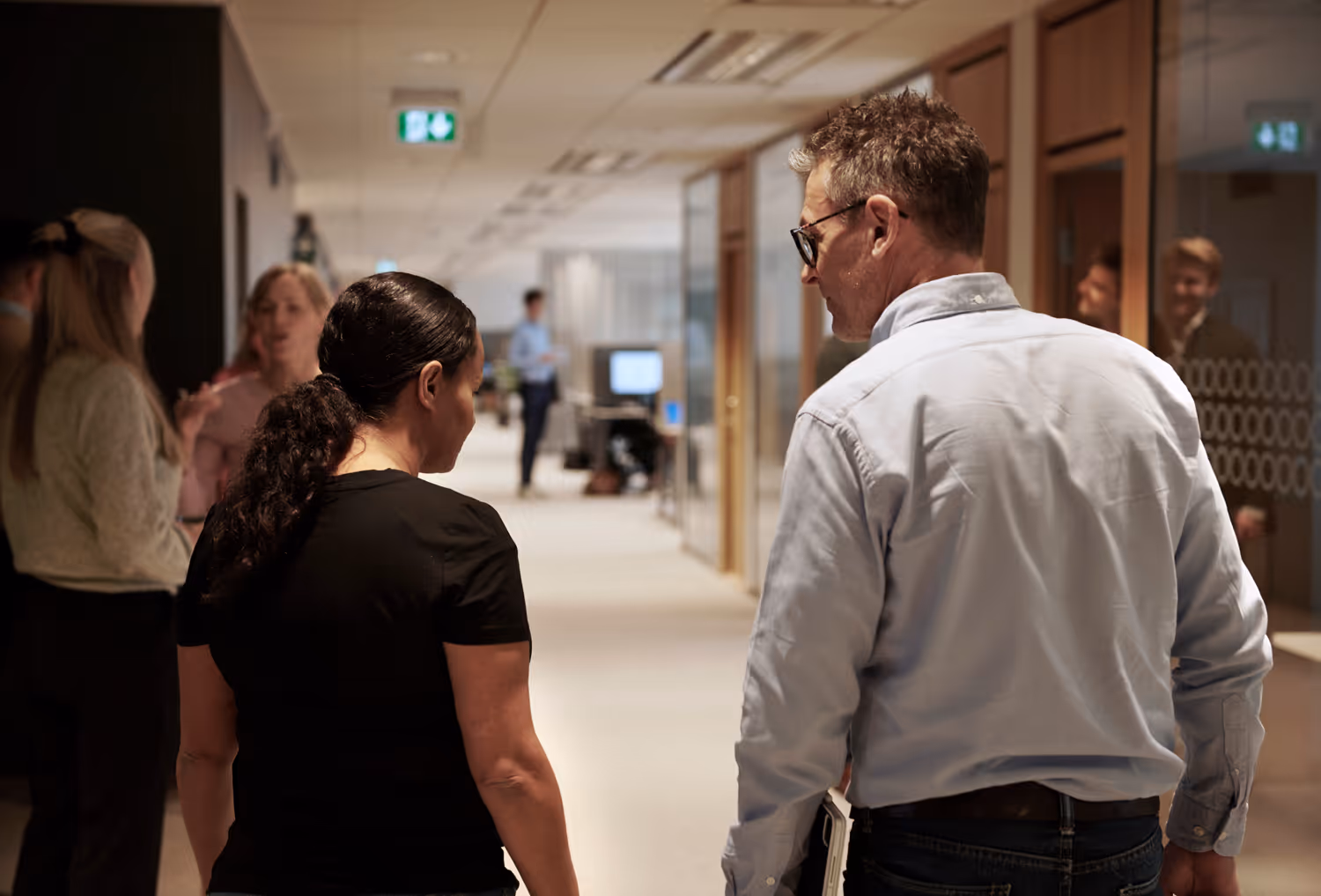 To personer går og snakker i en moderne kontorgang med flere kolleger i bakgrunnen.