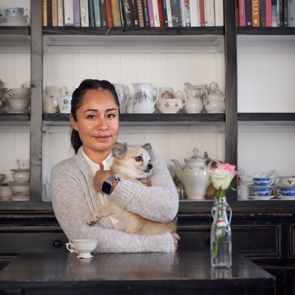 Kvinne i grå genser holder en liten hund foran et skap med servise og bøker.