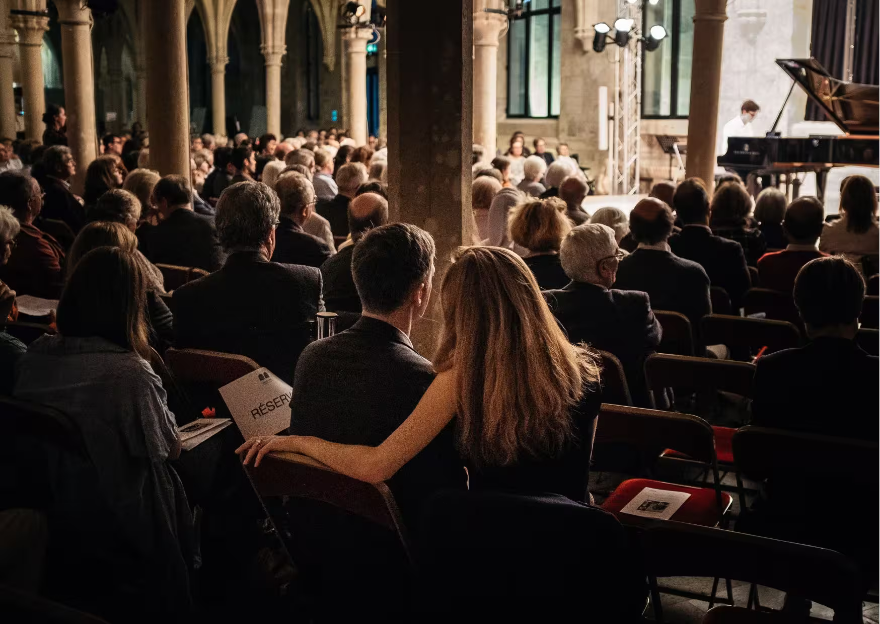 Un public assis dans un lieu avec de grandes arches, regardant un pianiste jouer sur scène, avec un couple au premier plan où la femme pose son bras autour de l'homme.