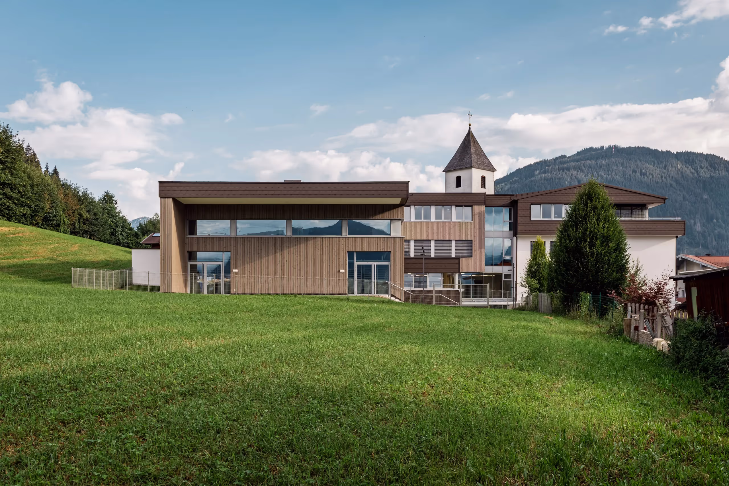 Aussenaufnahme der Volksschule Eben im Pongau, Holzfassade, geradlinige Architektur und Kirchturm im Hintergrund, saftige Weide im Vordergrund