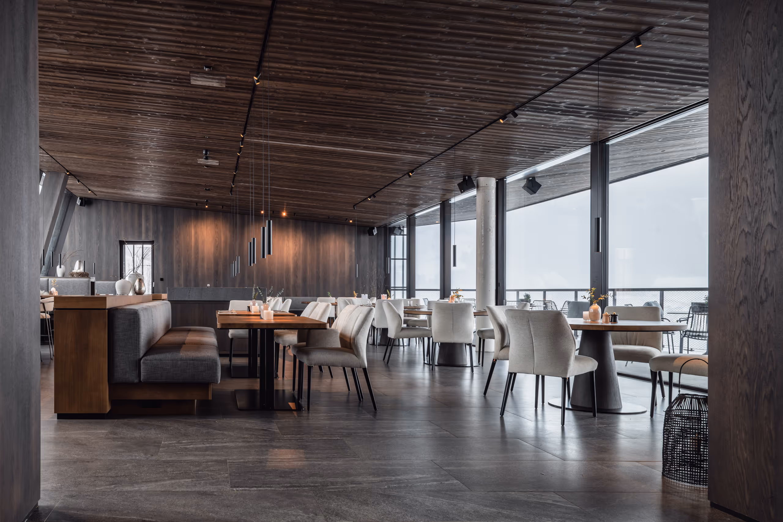 Großer Speisesaal des Hotel "Haven" im Alpendorf mit Designerstühlen, Holzverkleidung an den Wänden und der Decke, und riesigen Panoramafenstern mit Blick auf eine idyllische Winterlandschaft