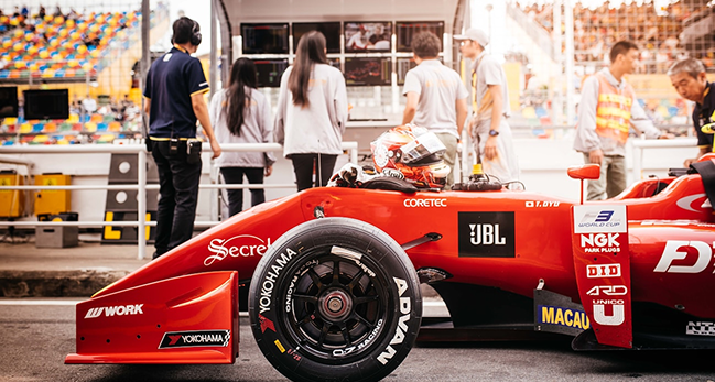 Red open-wheel race car with Yokohama tires in a garage area with team members and engineers in the background.