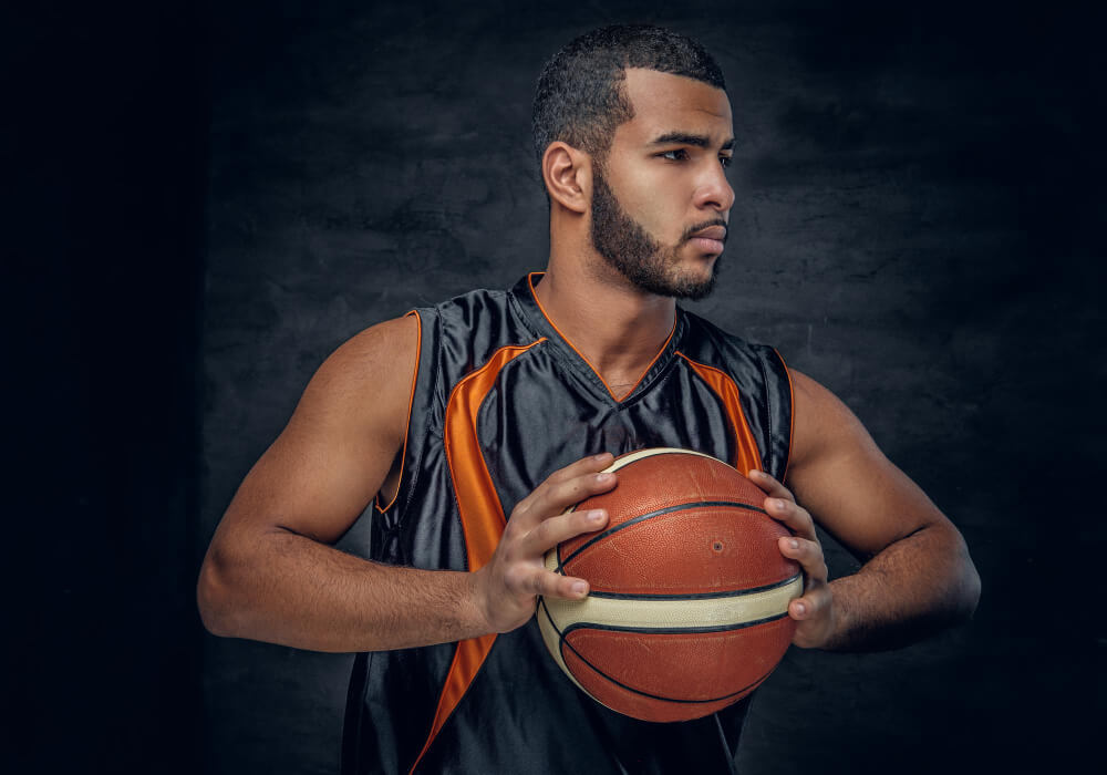 Young man in a black and orange basketball jersey holding a basketball with both hands, looking to the side against a dark background.