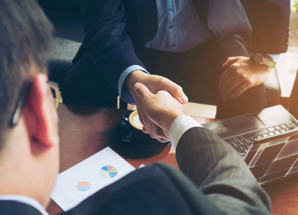 Two businessmen in suits shaking hands over a desk with a laptop, coffee cup, and documents with charts.