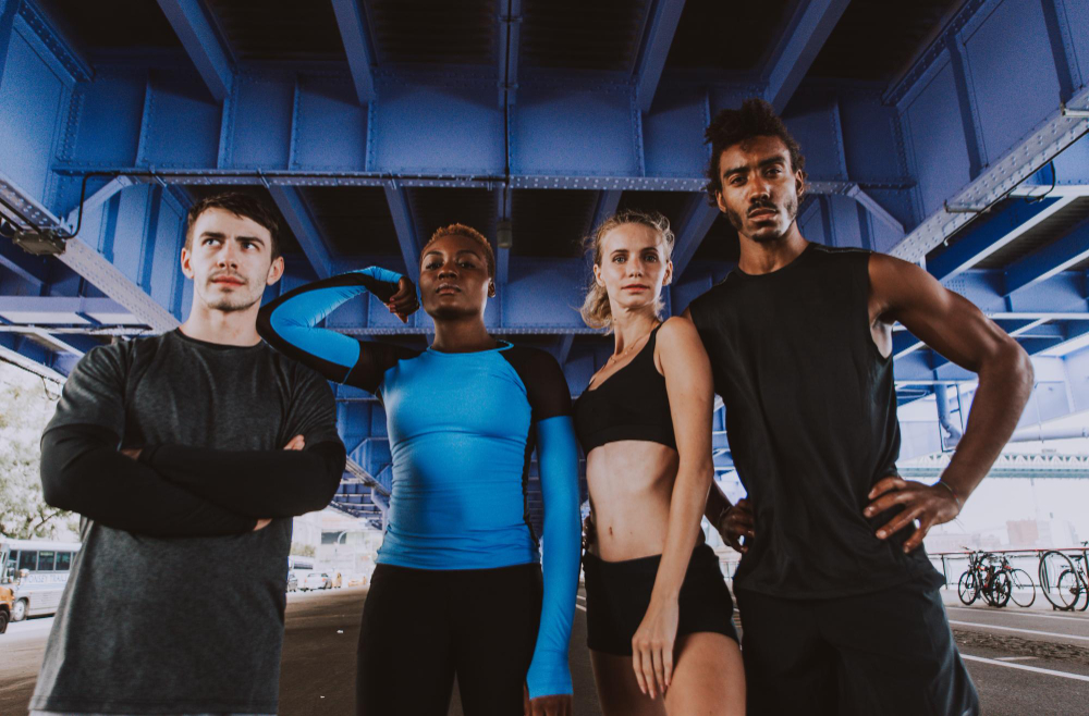 Four diverse athletes standing confidently under a blue metal bridge, dressed in sportswear.