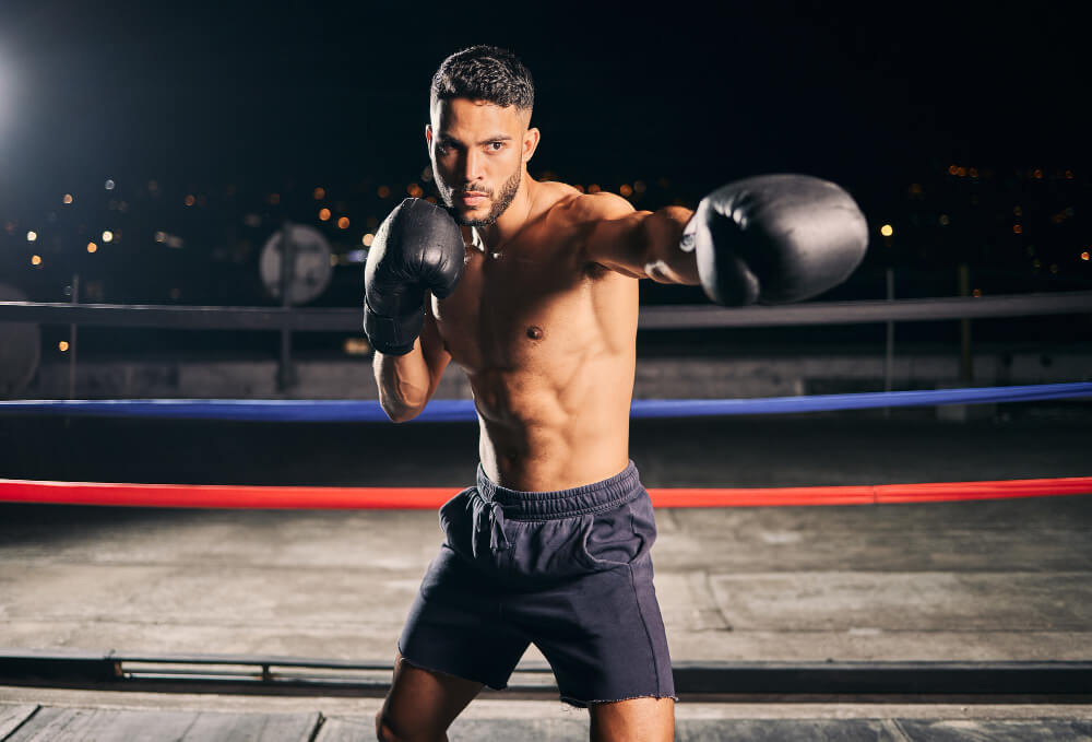 Shirtless male boxer wearing black gloves and dark shorts throwing a punch in a boxing ring at night.