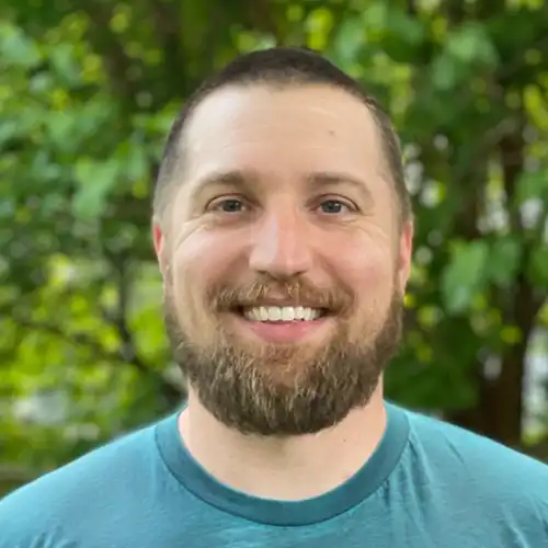 Smiling man with short brown hair and beard wearing a teal shirt outdoors with green foliage background.