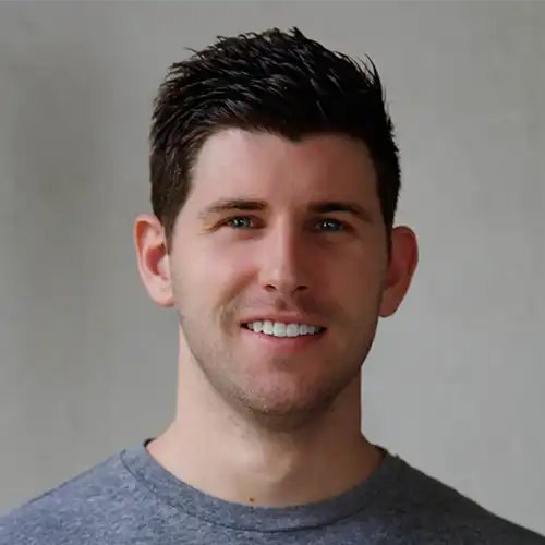 Young man with short dark hair and blue eyes smiling against a plain gray background, wearing a gray crew-neck shirt.