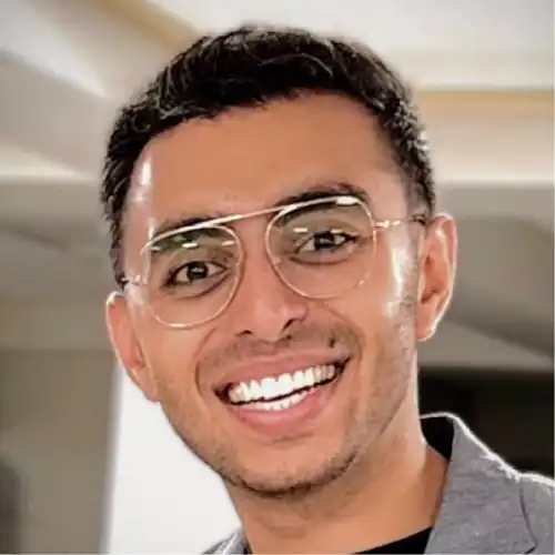 Smiling man with short dark hair and glasses wearing a gray jacket indoors.