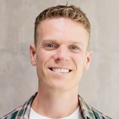 Smiling man with curly short hair wearing a plaid shirt over a white t-shirt against a plain background.