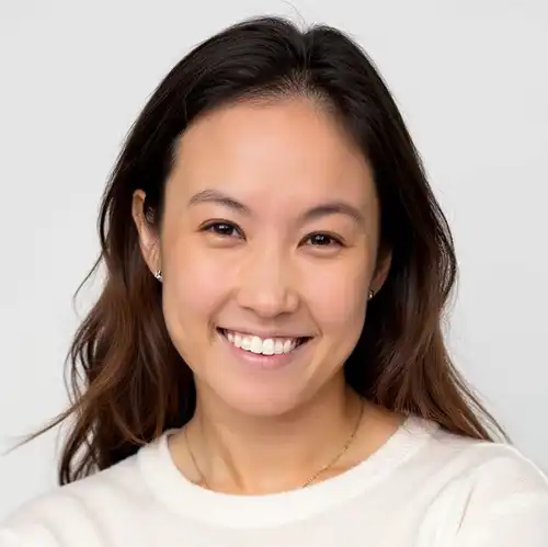 Smiling woman with long dark hair wearing a white sweater against a plain light background.