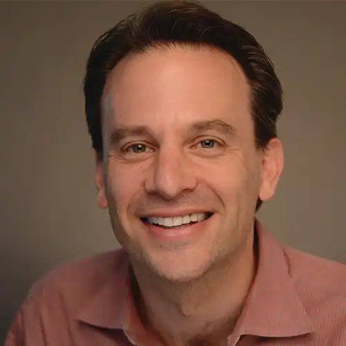 Smiling man with dark hair wearing a reddish shirt against a neutral background.