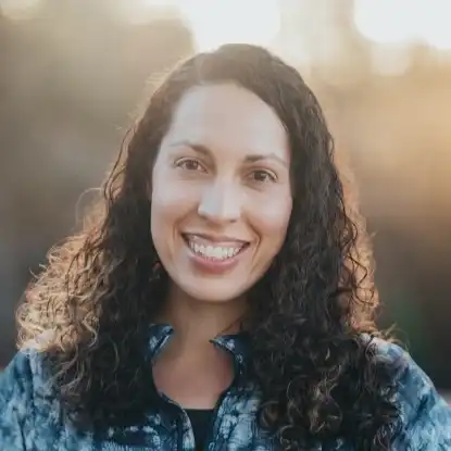 Smiling woman with long curly hair wearing a blue patterned jacket with sunlight in the background.