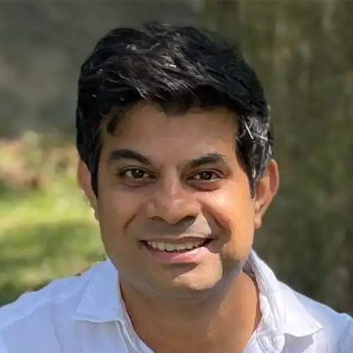 Smiling man with dark hair wearing a white shirt outdoors with greenery in the background.