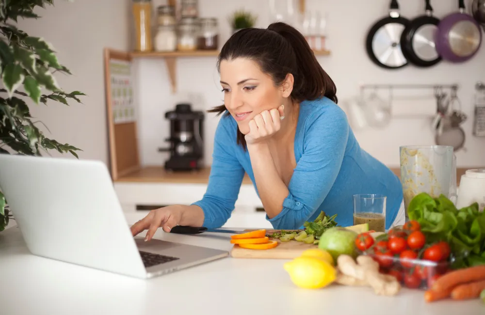Woman Reading Laptop in Kitchen. Vegetables next to her.