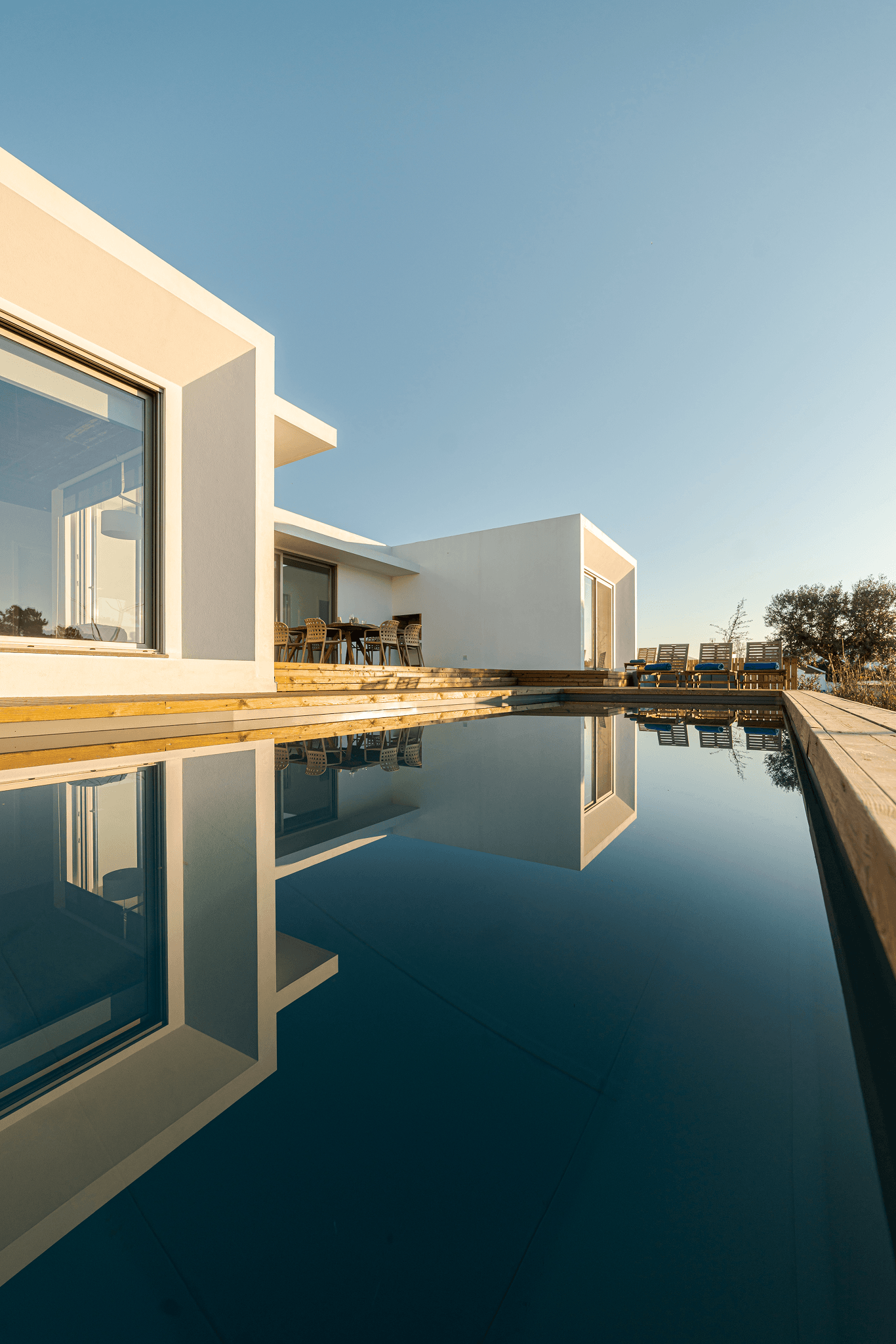 Modern white house with large windows and wooden deck reflected in a calm swimming pool under clear sky.