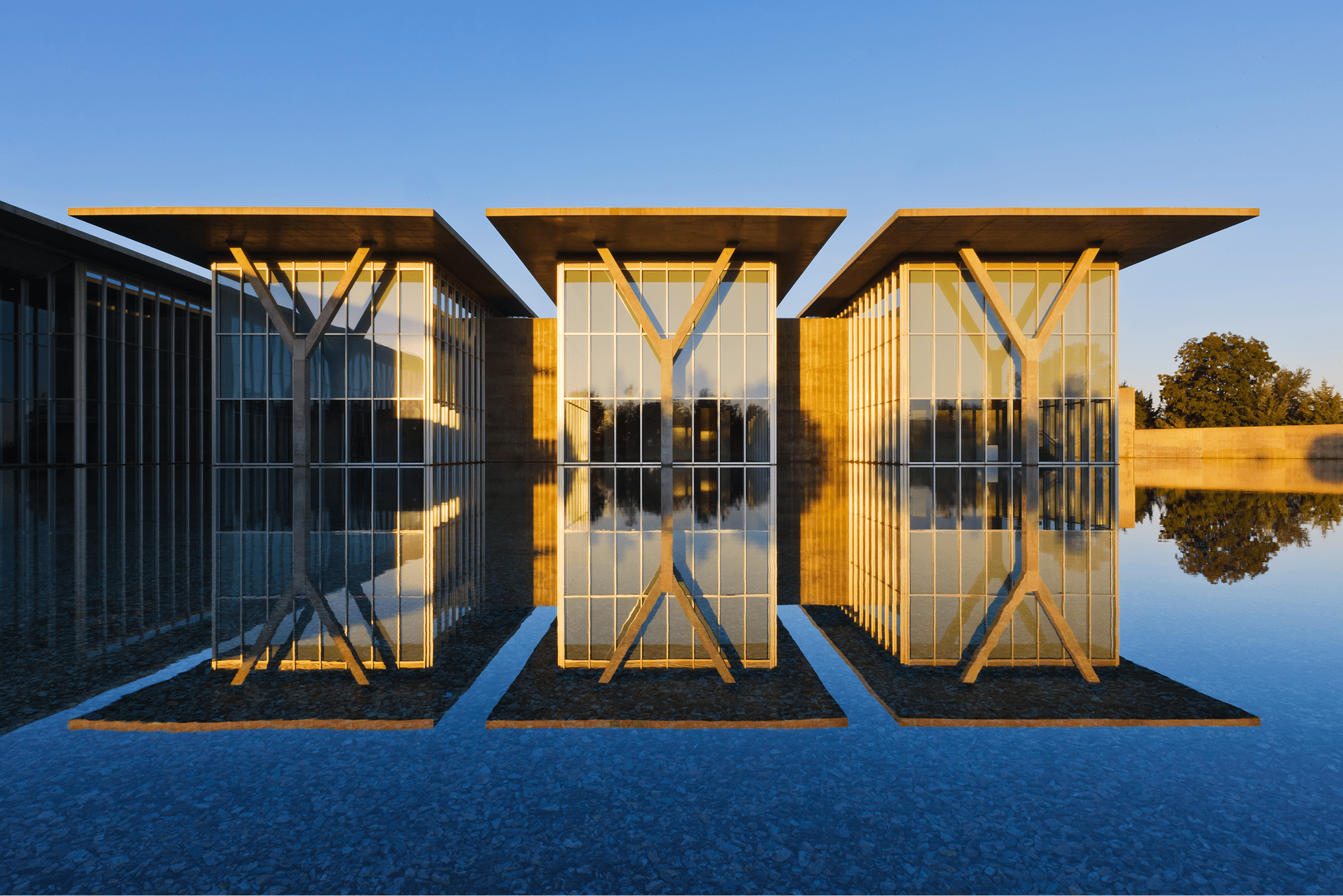 Three modern glass buildings with flat roofs reflected in calm water at sunset.