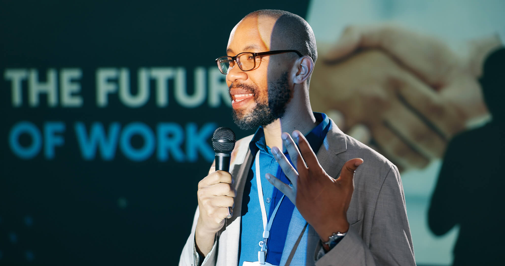 Man wearing glasses and suit jacket speaking into a microphone in front of a presentation screen.