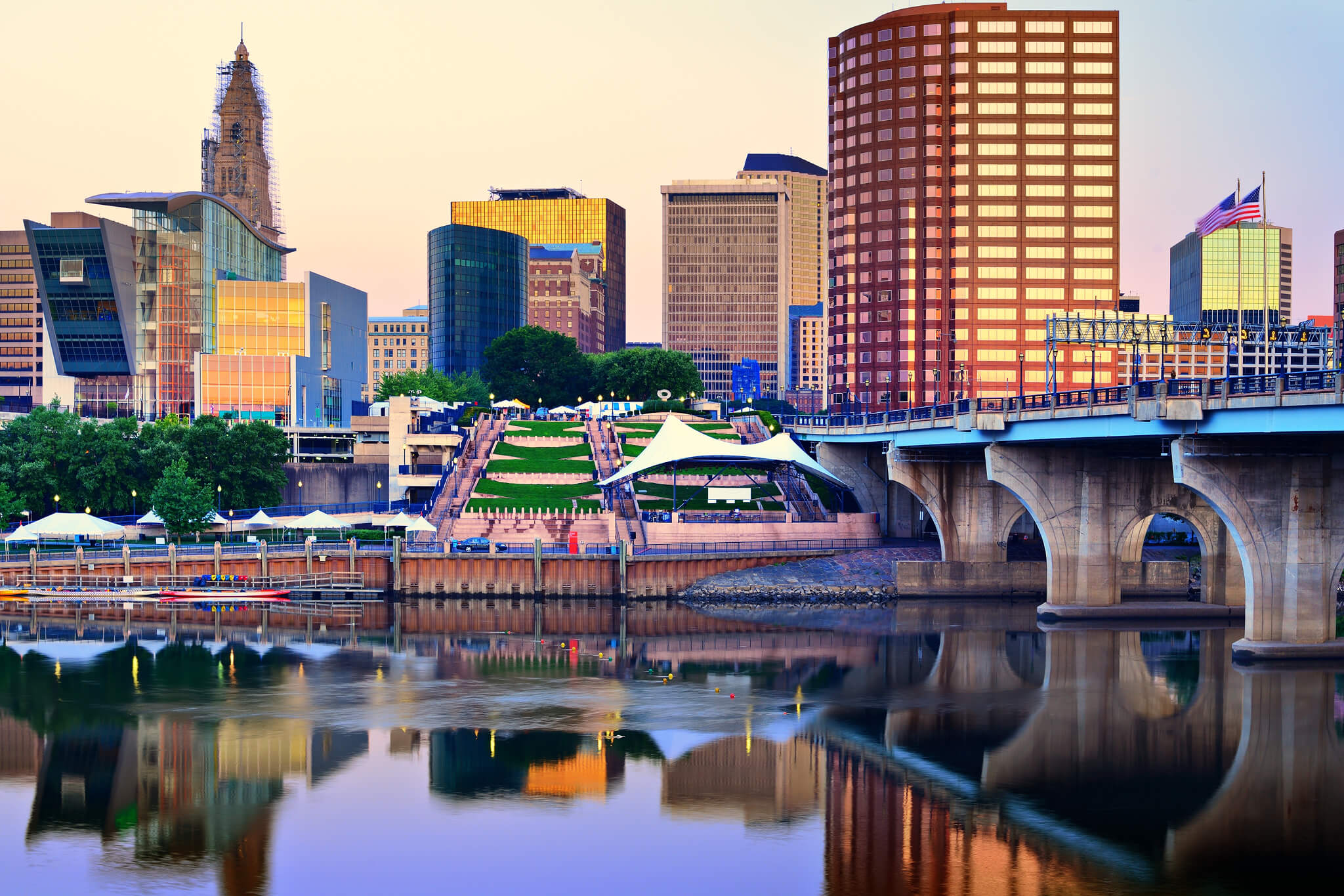 Downtown city skyline with modern buildings and a bridge reflected in calm river water at sunset.