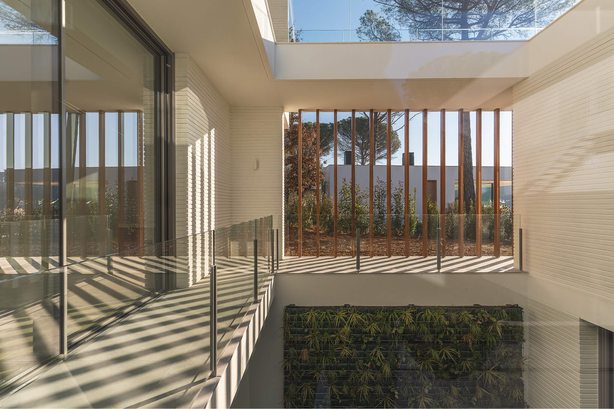 Modern balcony with glass railing, vertical wooden slats, white brick walls, and a green plant wall below.