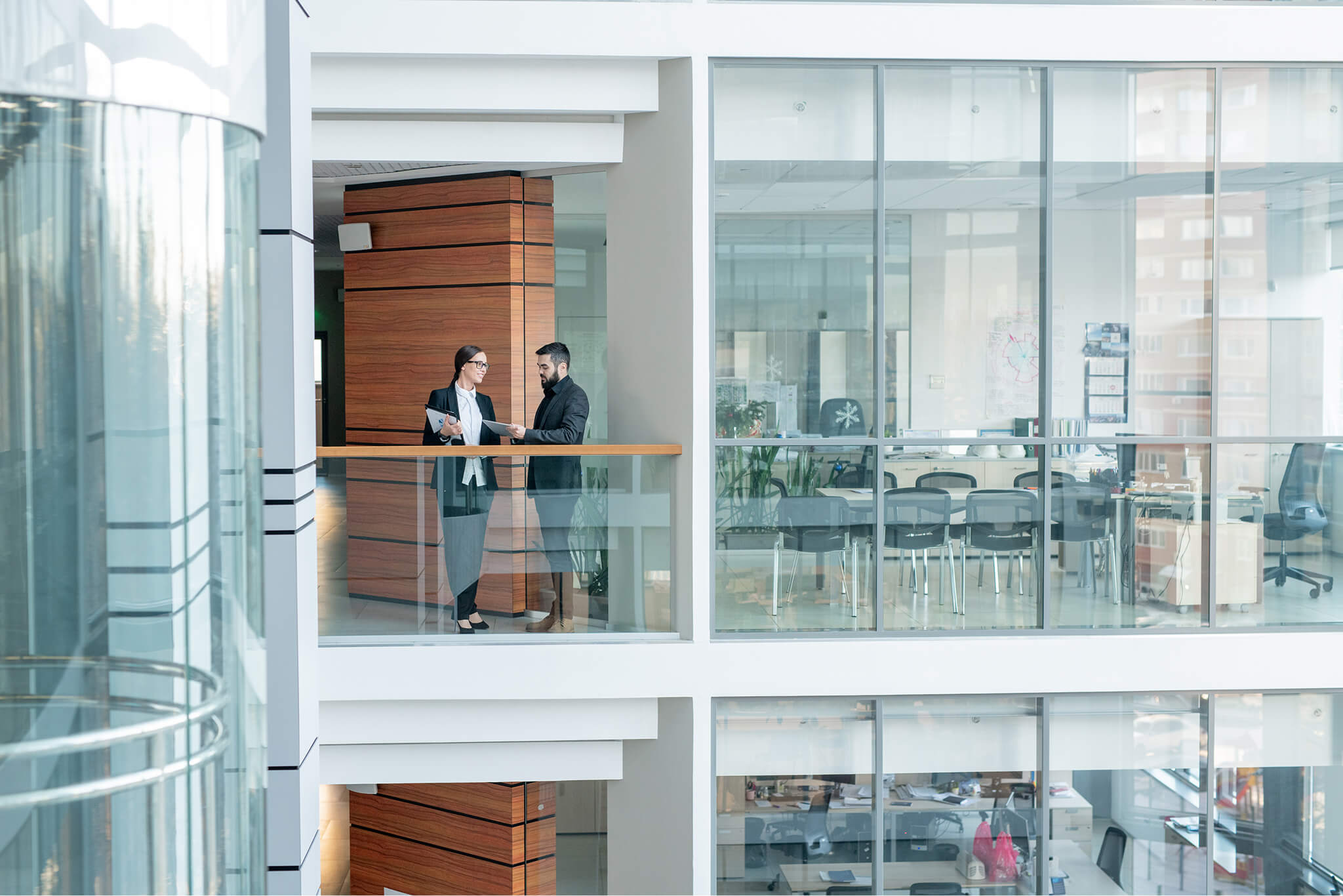 Two business professionals standing and discussing documents in a modern office building balcony area.