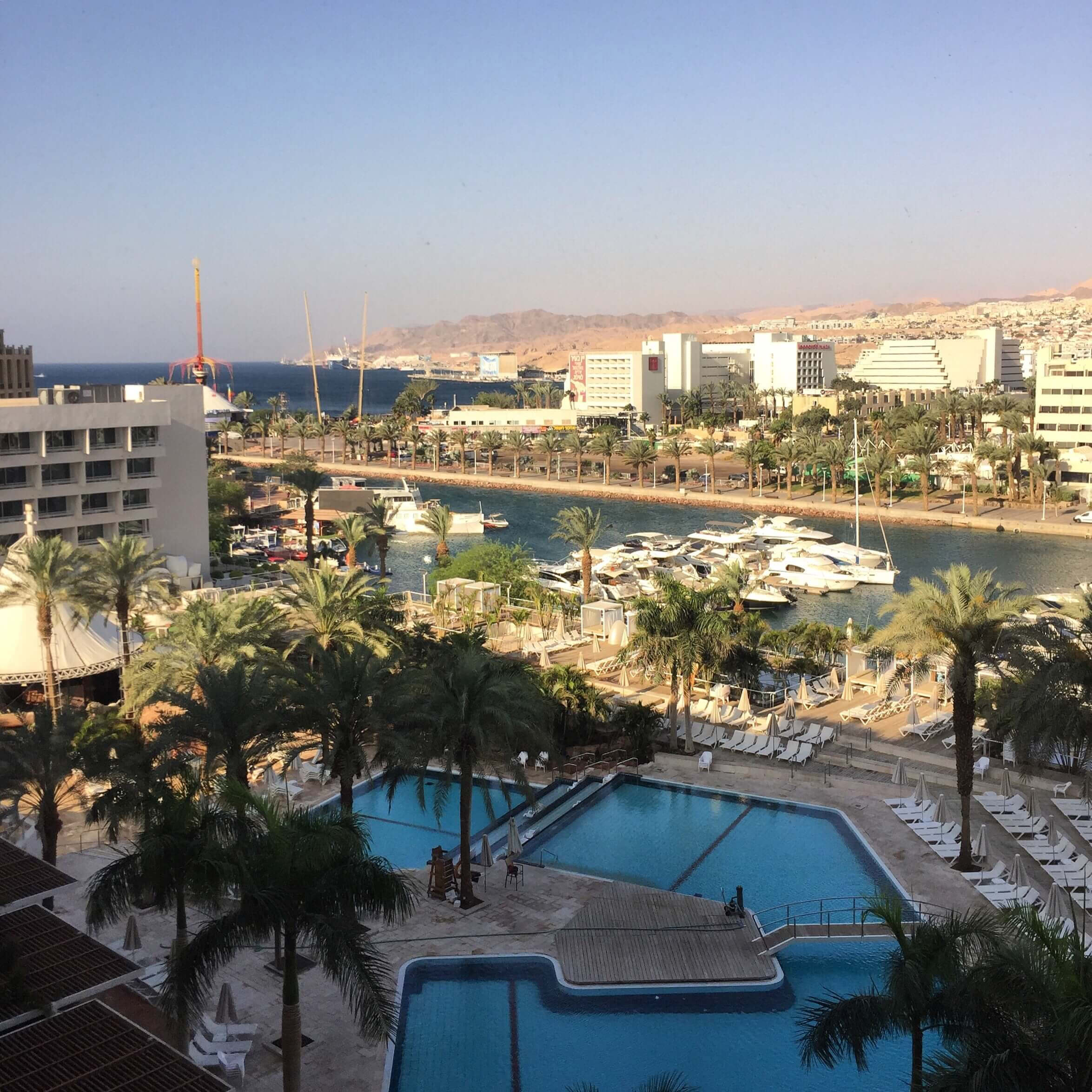View of resort pool, palm trees, marina with boats, waterfront hotels, and distant desert hills under a clear sky.