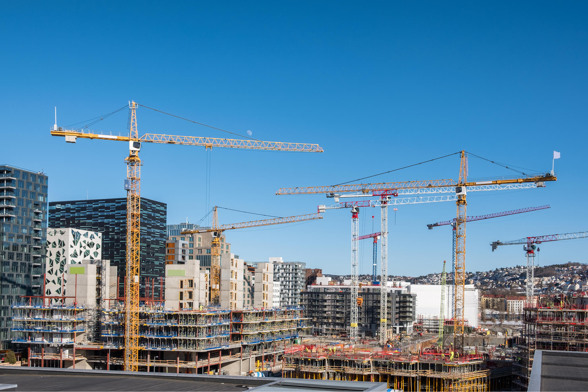 Multiple construction cranes over a large urban building site with partially built structures and clear blue sky.