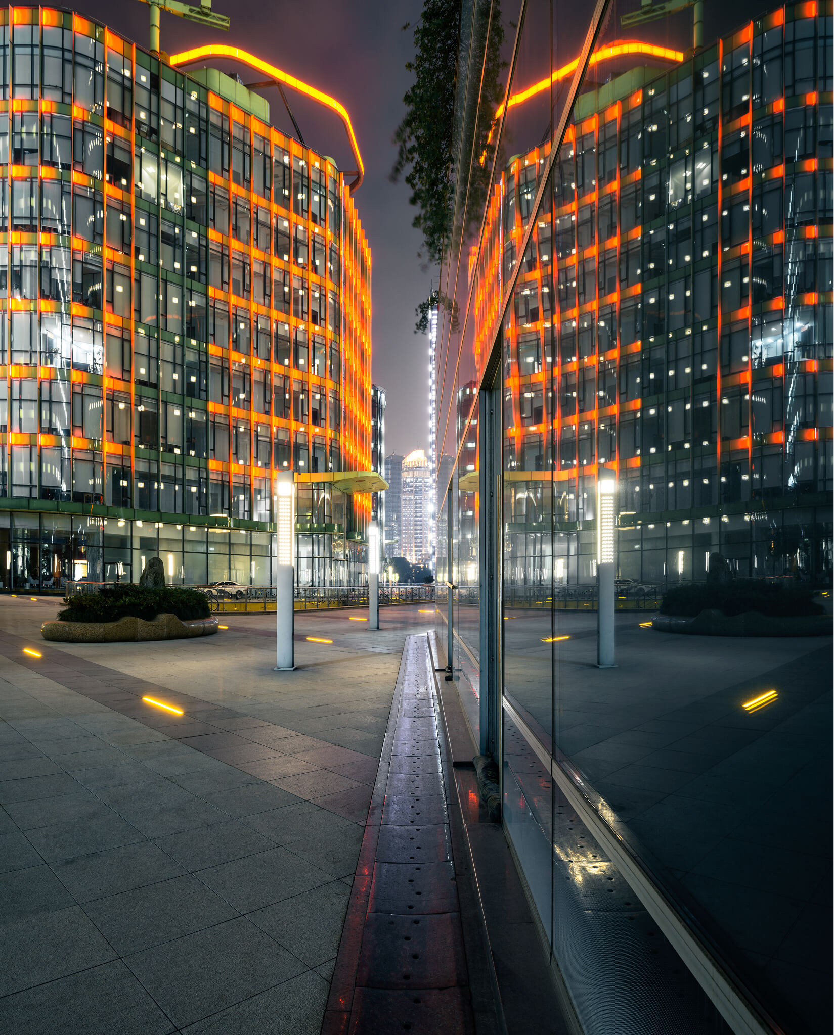 Modern glass building lit with orange lights reflected in adjacent glass wall at night.