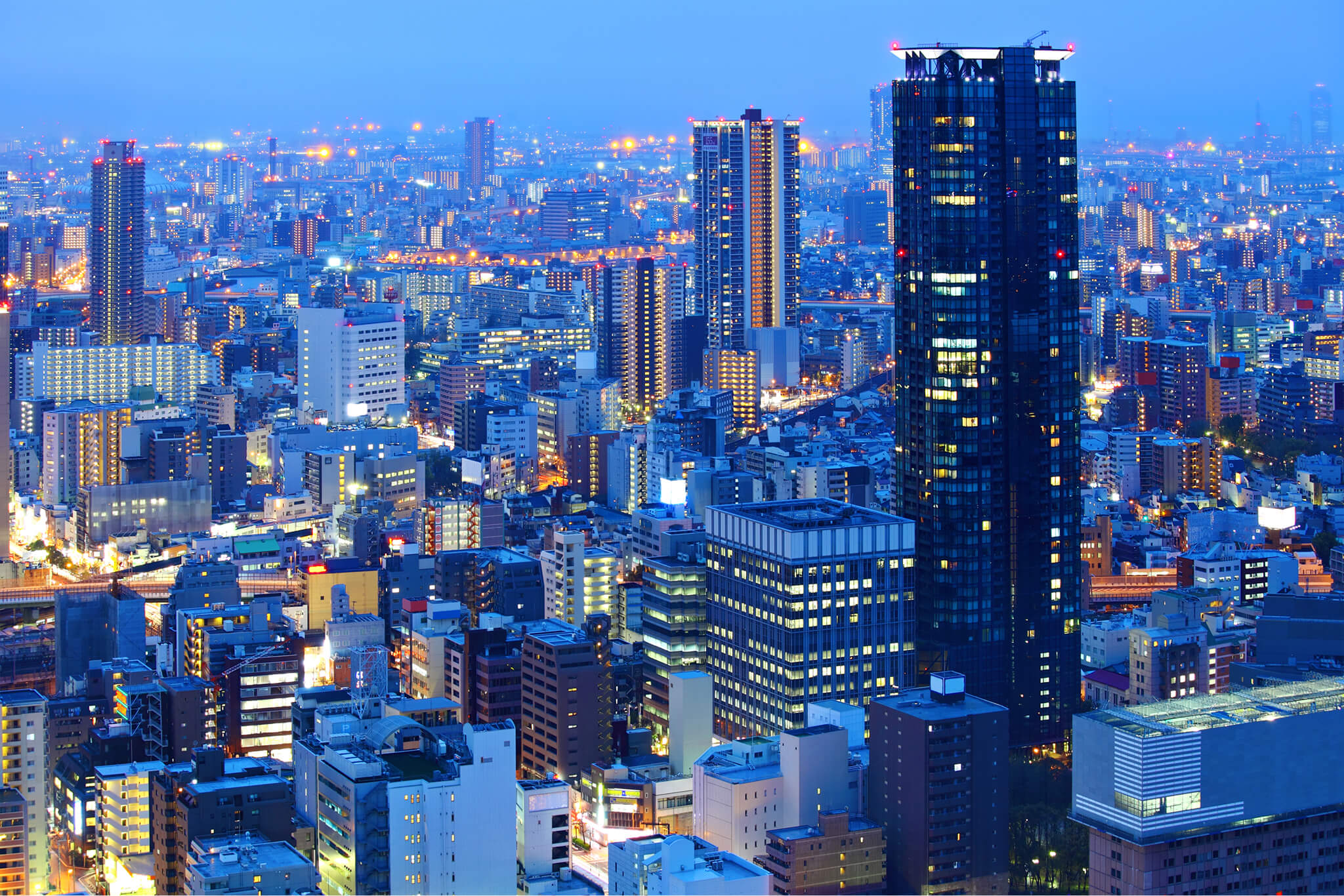 Cityscape at dusk with illuminated skyscrapers, high-rise buildings, and streetlights under a blue sky.