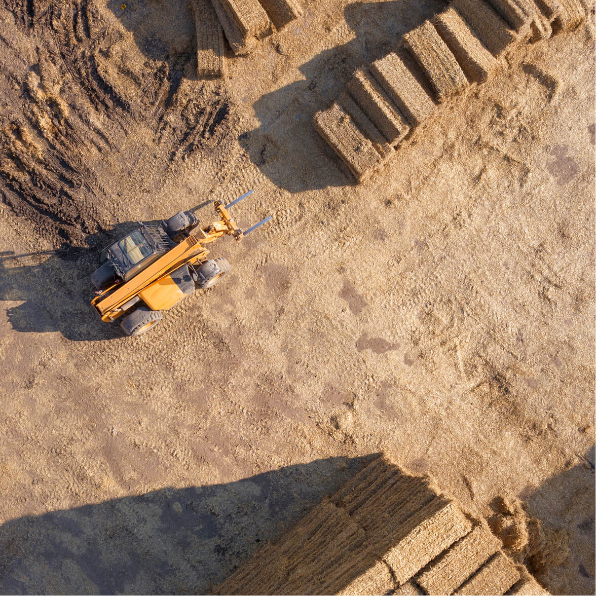 Aerial view of a yellow forklift on dirt near stacked rectangular hay bales casting shadows.