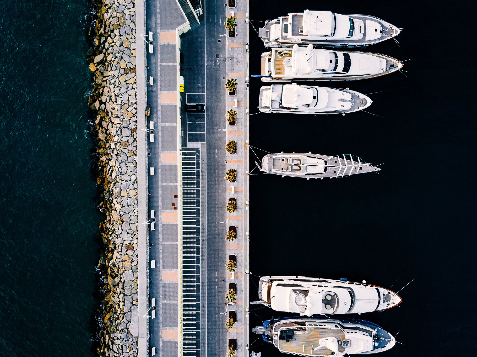 Aerial view of five white boats docked along a pier with a rocky seawall on one side.