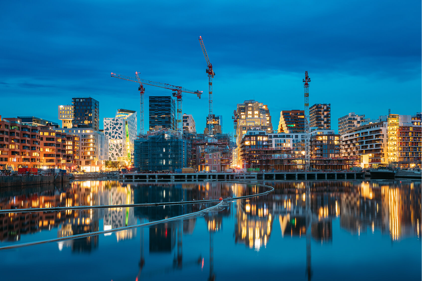 City skyline at dusk with modern buildings, construction cranes, and reflections on calm water.